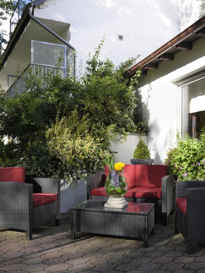 Gemütliche Sitzecke im Freien mit roten und grauen Möbeln. Üppige Pflanzen und ein blühender Topf sorgen für eine einladende Atmosphäre.