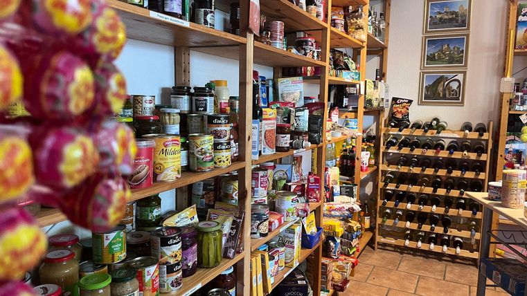 Een goed gesorteerde supermarkt met schappen vol potten, blikken en snacks. Op de achtergrond zijn ook wijnvaten te zien.