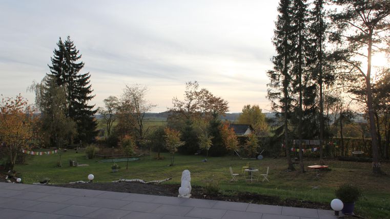 A peaceful garden landscape with autumn trees and a clear sky. In the foreground, seating and some garden ornaments are visible.