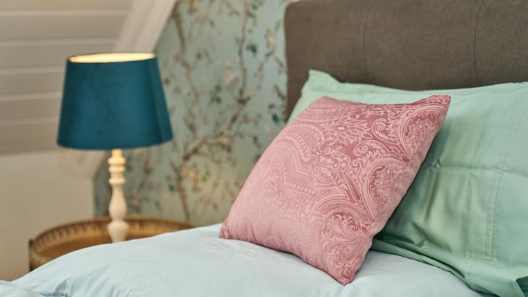 A cozy bedroom with a light blue bed and a pink pillow. A lamp with a green shade provides soft light.