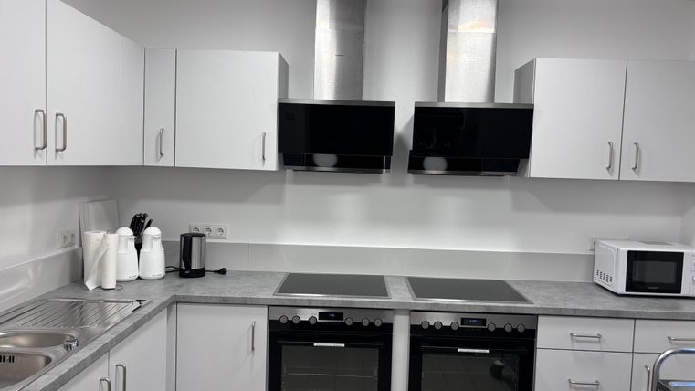 Eine moderne Küche mit weißen Schränken und einer grauen Arbeitsplatte. Sie ist ausgestattet mit zwei Kochfeldern, zwei Öfen und einem Mikrowellenofen.