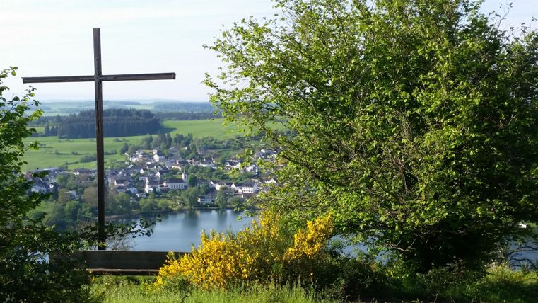 Ein Kreuz steht auf einem Hügel mit Blick auf ein kleines Dorf und einen Fluss. Umgeben von üppigem Grün und blühenden Sträuchern.