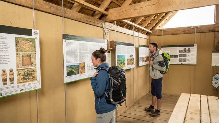 Zwei Personen mit Rucksäcken betrachten Informationstafeln in einem hölzernen Infopunkt. Die Tafeln zeigen archäologische Informationen.