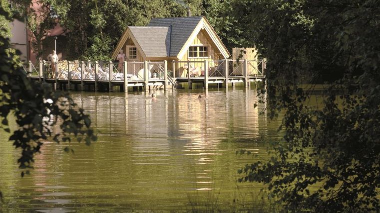 A cozy house by the water with a wooden terrace.Surrounded by trees, the building reflects in the calm lake.