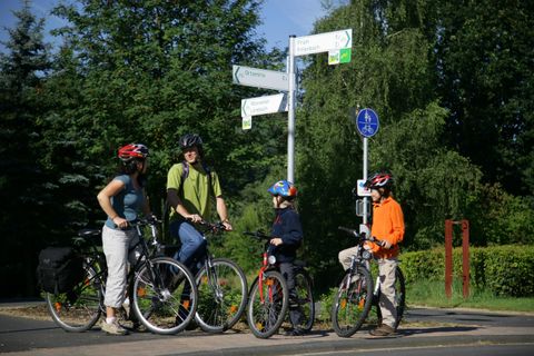 Eine Gruppe von Radfahrern steht an einem Wegweiser. Sie tragen Helme und bereiten sich auf den weiteren Weg vor.