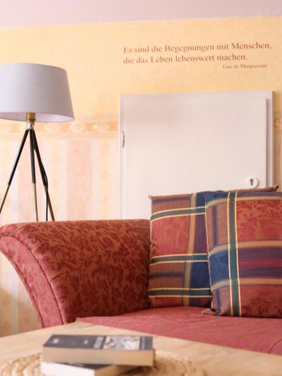 Ein gemütliches Wohnzimmer mit einer roten Couch und bunten Kissen. Neben der Couch steht eine moderne Lampe und an der Wand ist ein Zitat zu sehen.