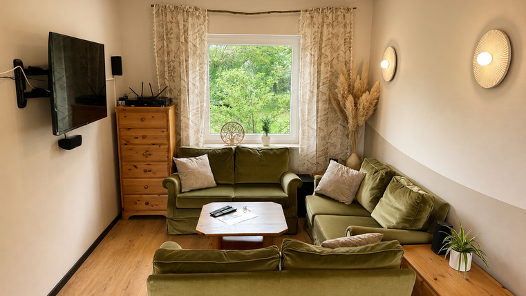 Ein gemütliches Wohnzimmer mit zwei grünen Sofas und einem Holztisch. Durch das Fenster sieht man ins Freie, umgeben von Pflanzen und natürlichem Licht.