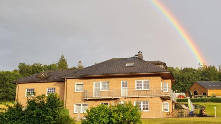 Ein zweistöckiges Haus mit einem Balkon und großen Fenstern steht in einer grünen Umgebung. Im Hintergrund gibt es einen Regenbogen, der den Himmel schmückt.