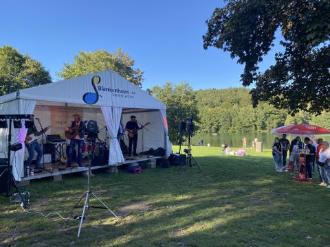 A band is playing live under a tent by the shore of a lake. In the background, there are people relaxing and enjoying nature.