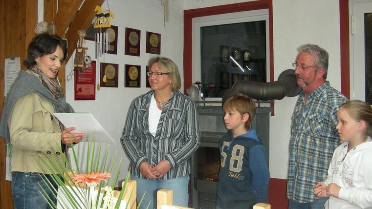 A small group of four people is standing in a cozy room. A woman is reading a document aloud while the others are listening attentively.