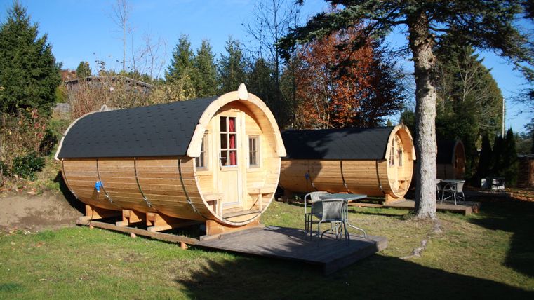 Zwei gemütliche Holzfass-Unterkünfte stehen in einem schönen Garten. Umgeben von Bäumen bietet der Bereich eine entspannende Atmosphäre.