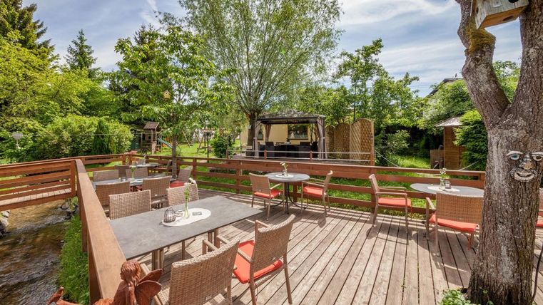 Eine gemütliche Terrasse mit Stühlen und Tischen aus Holz. Umgeben von grünen Bäumen und einem kleinen Bach.