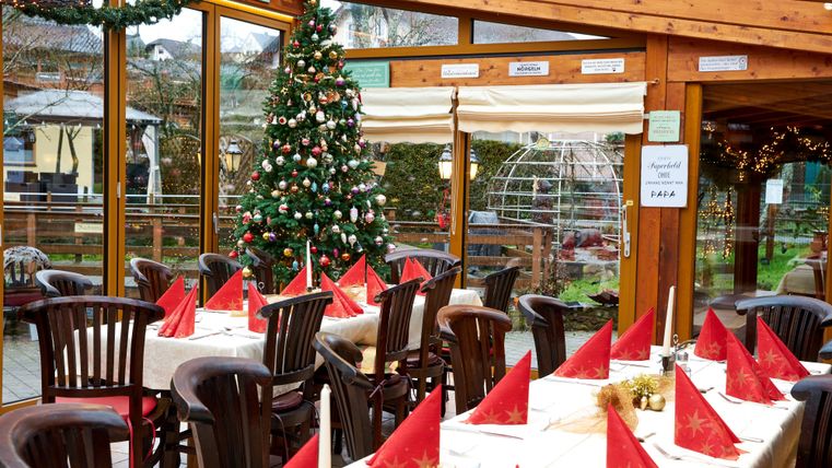 Ein festlich dekorierter Essbereich mit Holzstühlen und weißen Tischen. Im Hintergrund steht ein Weihnachtsbaum, der den Raum gemütlich gestaltet.