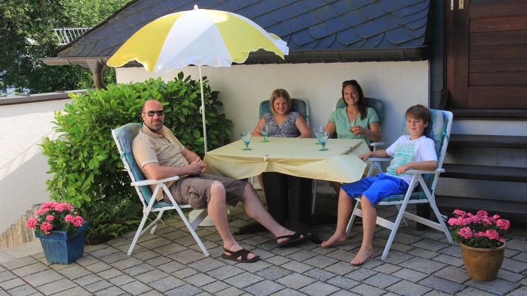 Eine Gruppe von vier Personen sitzt entspannt an einem Tisch unter einem Sonnenschirm. Um sie herum stehen Blumen in Töpfen, und die Stimmung ist freundlich und gemütlich.