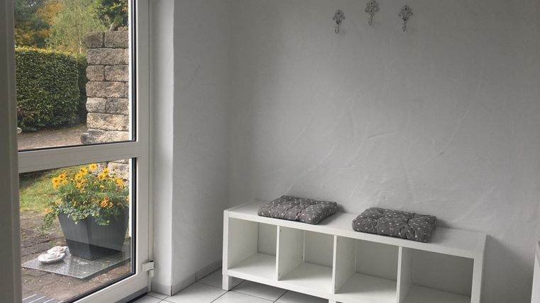 A bright room with white walls and tiles. There is an open door to a garden and a simple seating area with cushions.