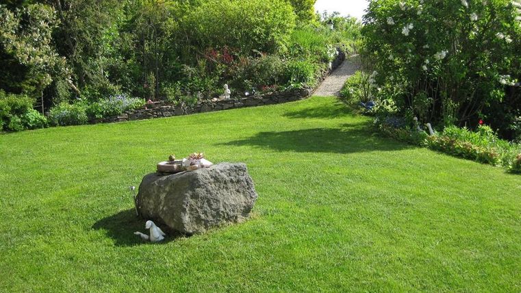 Een verzorgde tuin met groen gras en een grote steen in het midden. Op de achtergrond zijn kleurrijke struiken en een pad zichtbaar.
