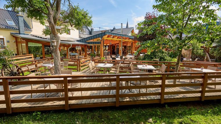A sunny terrace with wooden furniture and trees, surrounded by a wooden fence, in front of a building with large windows.