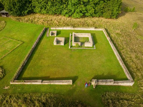 Eine archäologische Stätte mit grasbewachsenen Ruinen und Steinmauern. Zwei Personen stehen auf dem Gelände und betrachten die Überreste.