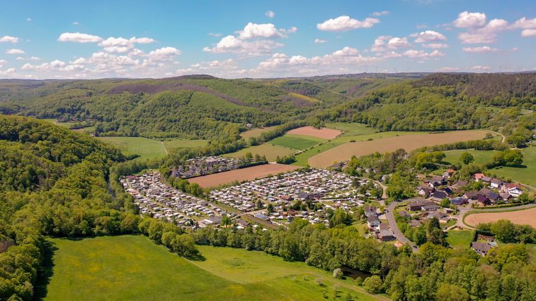 An aerial view of a campsite surrounded by green hills and meadows. The sky is clear with a few clouds.