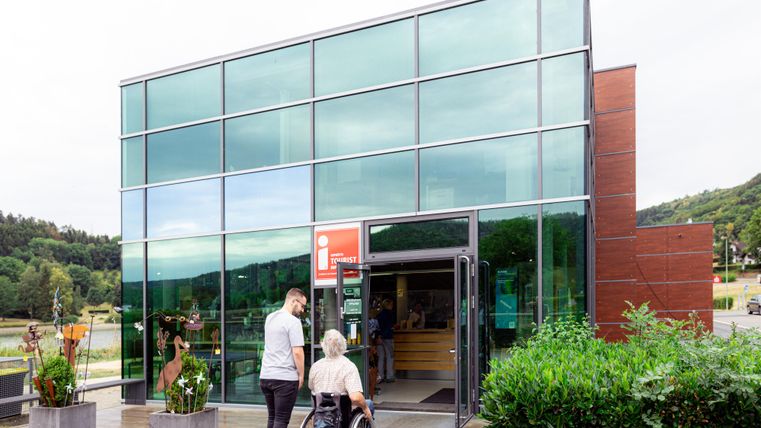 Ein modernes Gebäude mit großen Glasfenstern und einem Eingang. Eine Person im Rollstuhl wird von jemandem begeleitet.