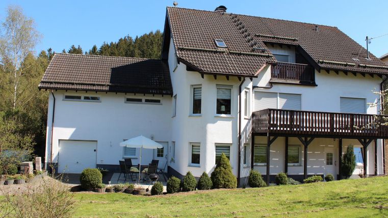 A modern house with a large garden and porch. The sun is shining and the surroundings are green and inviting.