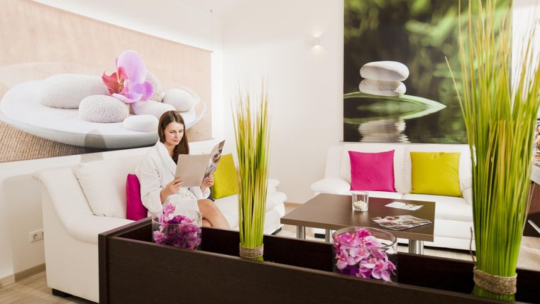 Een moderne ontspanningsruimte met een witte bank en kleurrijke kussens. Een vrouw in een badjas leest in een menu, omringd door groene planten en decoratieve stenen.