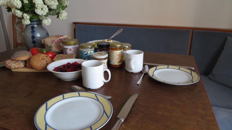 Ein schön gedeckter Tisch mit zwei Tellern, Tassen und verschiedenen Marmeladen. Im Hintergrund sind frische Brötchen und eine Blumenvase zu sehen.