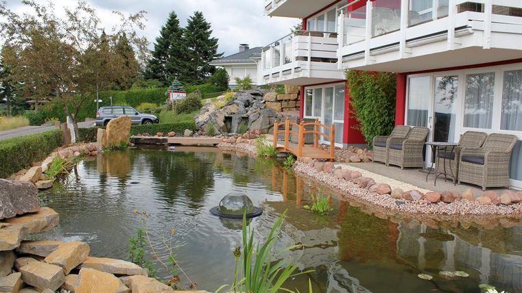 Ein schöner Teich mit Pflanzen und Steinen, umgeben von einem modernen Gebäude. Auf der Terrasse stehen gemütliche Stühle, ideal zum Entspannen.