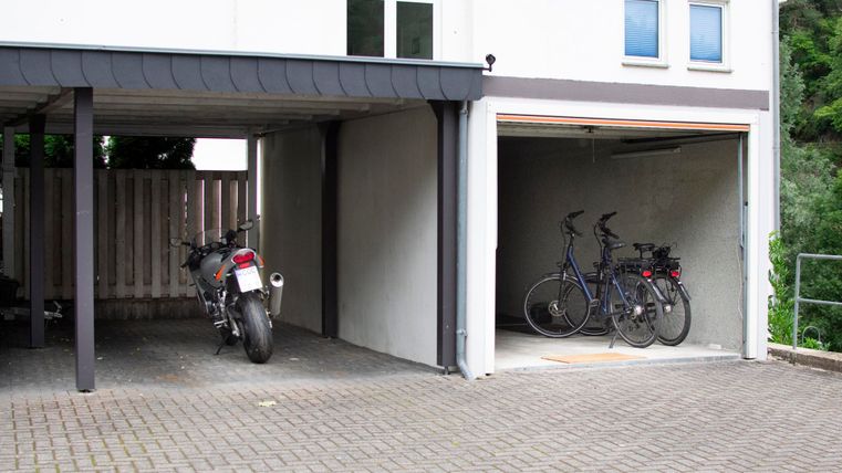Eine zweigeteilte Garage mit einem Motorrad auf der linken Seite und zwei Fahrrädern auf der rechten Seite. Der Boden ist mit Pflastersteinen ausgelegt und es gibt Fenster in der oberen Etage.