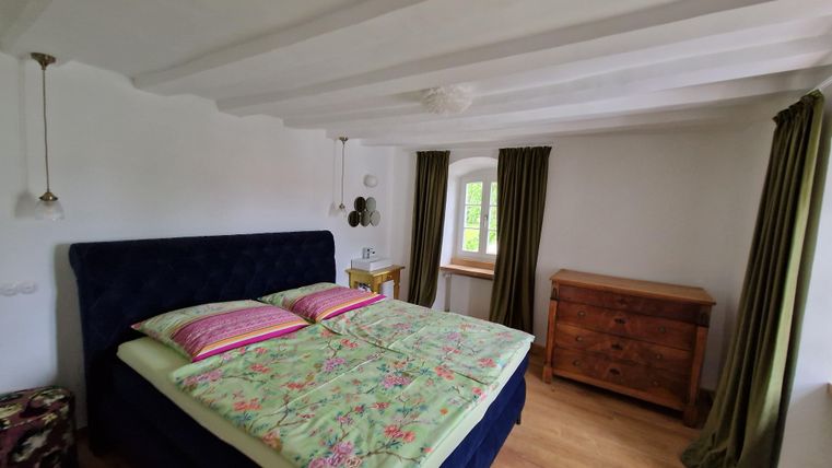 A cozy bedroom with a large bed and colorful pillows. There is a wooden wardrobe and green curtains that let in light.