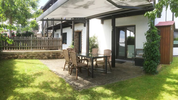 Ein gemütlicher Außenbereich mit einem Tisch und Stühlen unter einem Sonnenschutz. Umgeben von grünem Rasen und einem Sichtschutzzaun.