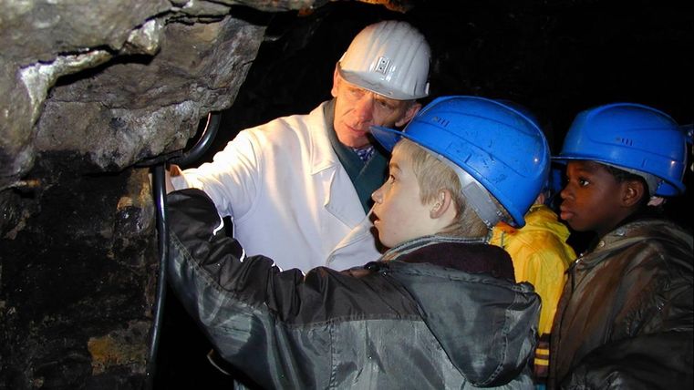 A group of children and an adult in a mine. They are wearing safety helmets and are looking intently at an object.