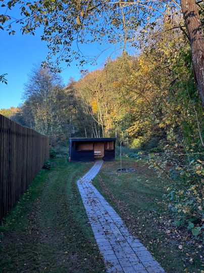 Ein ruhiger Weg führt zu einer kleinen Hütte inmitten von Bäumen und herbstlichen Blättern. Die Umgebung ist grün und friedlich, ideal für einen entspannten Aufenthalt in der Natur.