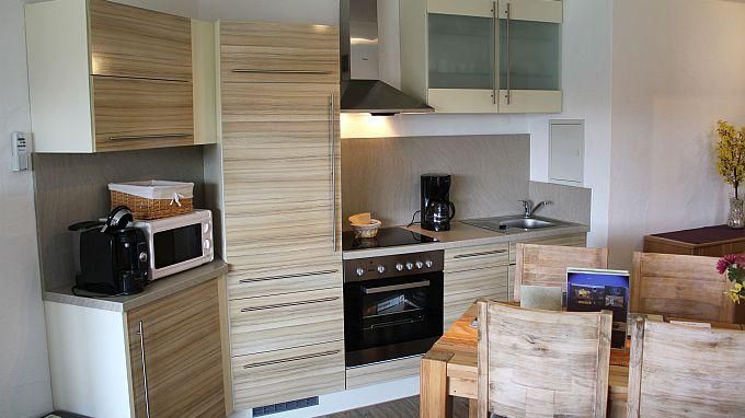 Eine moderne Küche mit hellen Holzschränken und Arbeitsflächen. Auf der linken Seite stehen ein Kühlschrank, eine Mikrowelle und eine Kaffeemaschine, während der Essbereich aus einem Holztisch mit Stühlen besteht.