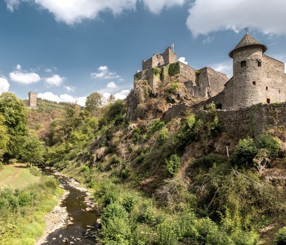 Die Manderscheider Burgen thronen auf einem Hügel inmitten einer grünen Landschaft, umgeben von Bäumen und einem kleinen Fluss unter blauem Himmel., © Dominik Ketz