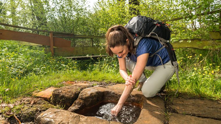 A woman with a backpack is kneeling at a small spring in the forest and washing her hands. In the background, a wooden bridge can be seen.