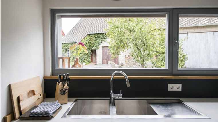 Eine moderne Küche mit einem Edelstahl-Spülbecken und Küchenutensilien. Durch das Fenster sieht man einen grünen Garten und ein charmantes Gebäude.