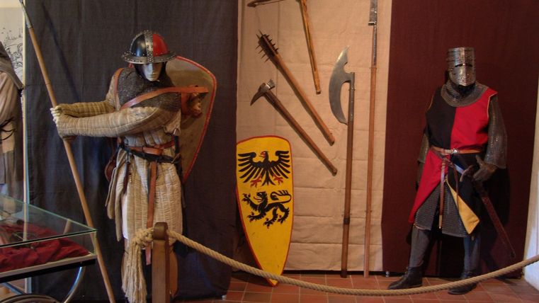 A medieval museum with two figures in armor. In the background, various weapons and a shield can be seen.