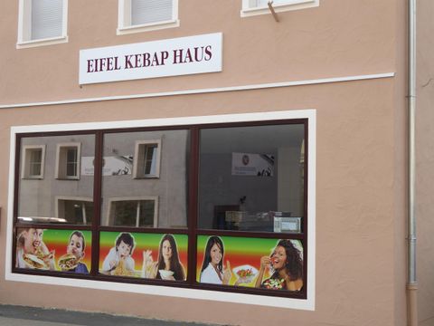 Ein Restaurant namens "Eifel Kebap Haus" mit großen Fenstern. Die Fassade zeigt Bilder von verschiedenen Gerichten und fröhlichen Menschen.