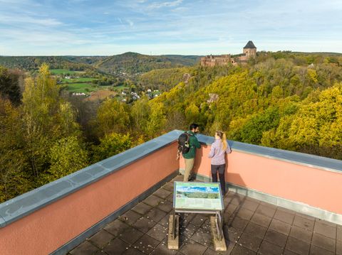 Een uitzicht vanaf een uitkijkpunt met twee personen die op het omringende landschap kijken. Op de achtergrond is een kasteelruïne op een heuvel te zien, omringd door kleurrijke bomen.