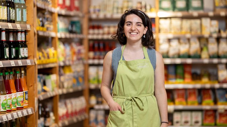 A friendly saleswoman in a store stands in front of shelves with groceries. She is wearing a green apron and smiling.