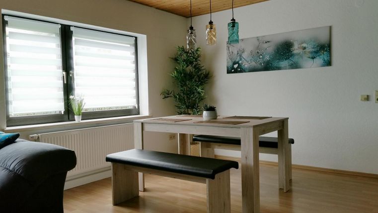 A modern dining room with a light wooden table and benches. Large windows provide plenty of natural light and a beautiful wall decoration complements the space.