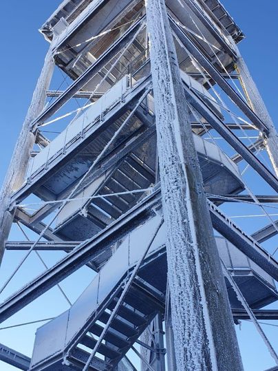 Ein hoher Aussichtsturm aus Metall, bedeckt mit Schnee und Eis, ragt in den blauen Himmel. Die Treppen führen spiralförmig nach oben.