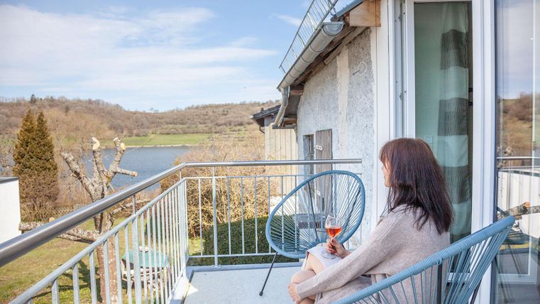 Eine Frau sitzt auf einem Balkon und genießt einen Drink. Im Hintergrund sind eine malerische Landschaft und ein Wasserlauf zu sehen.