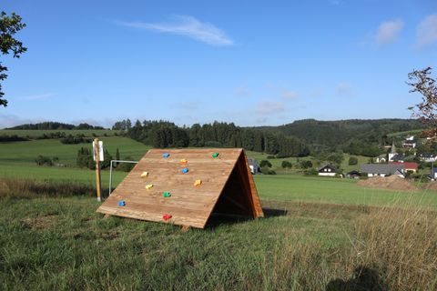 Ein Klettergerüst aus Holz steht auf einem Feld mit grünen Wiesen im Hintergrund. Der Himmel ist klar und blau, ideal für Outdoor-Aktivitäten.