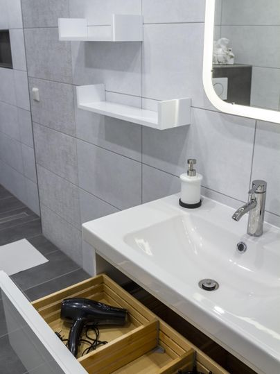 A modern bathroom with a white sink and shelf combination. In the open drawers are a hairdryer and other bathroom utensils.