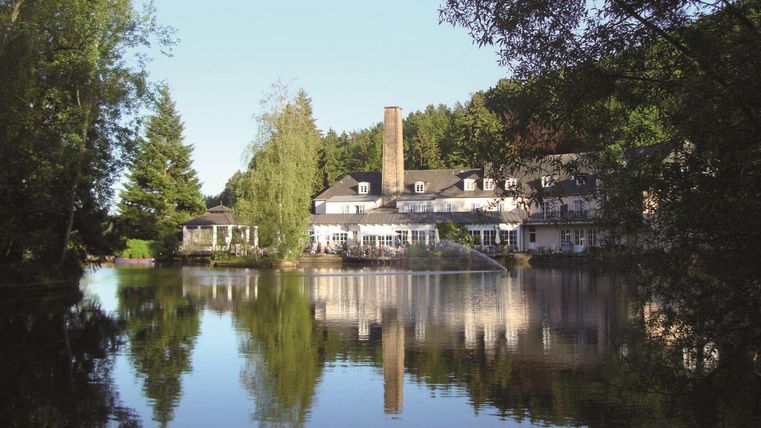 Ein ruhiges Hotel am Ufer eines Sees, umgeben von Bäumen. Die Reflexion des Gebäudes im Wasser schafft eine friedliche Atmosphäre.