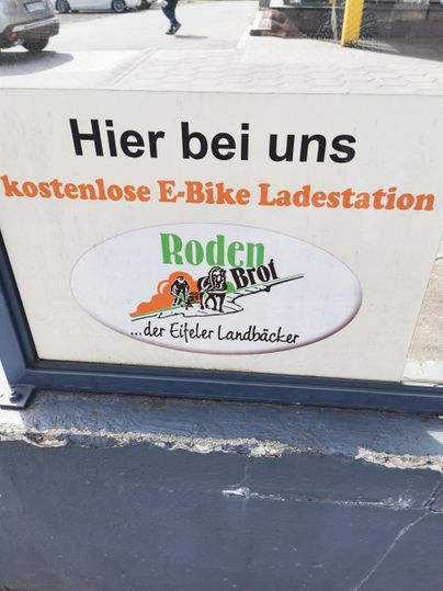 Eine Beschilderung mit der Aufschrift "Hier bei uns kostenlose E-Bike Ladestation" und dem Logo von "Roden & Brot". Der Hinweis ist in einer klaren und einladenden Schrift gestaltet.