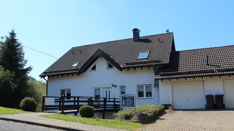A large white house with a tiled roof and a parking space in front of the door. In the background, there are trees and a clear blue sky.