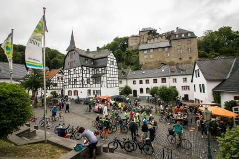 Een drukke markt met veel fietsers en historische gebouwen. Op de achtergrond is een kasteel te zien, omringd door bomen.